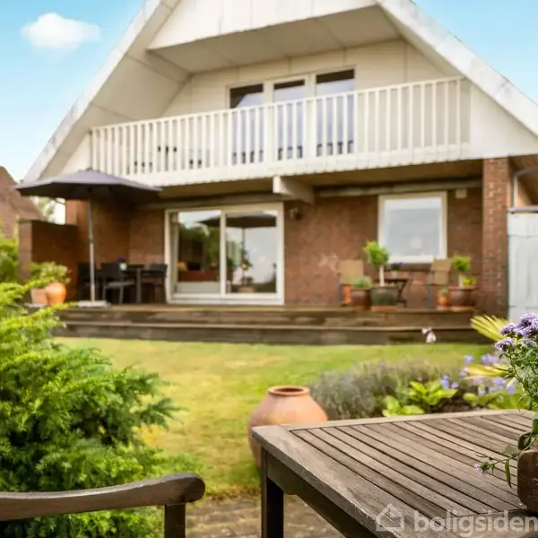 Hus med altan og terrasse set fra have. Forhave med bord, stole og blomster i forgrunden. Terrasse med parasol og potteplanter, omgivet af græsplæne og buske i et roligt kvarter.