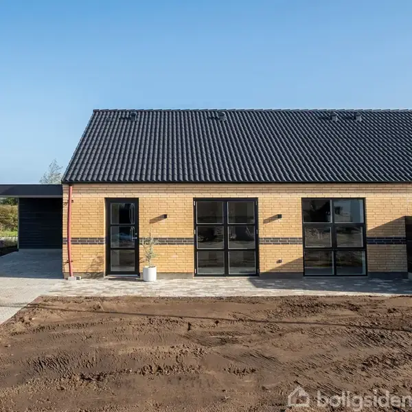 Et moderne murstenshus med store, sorte vinduer og en sort tag. Det står på en grund med ny jord foran og flisebelagt indkørsel til venstre. Klart, solrigt vejr.