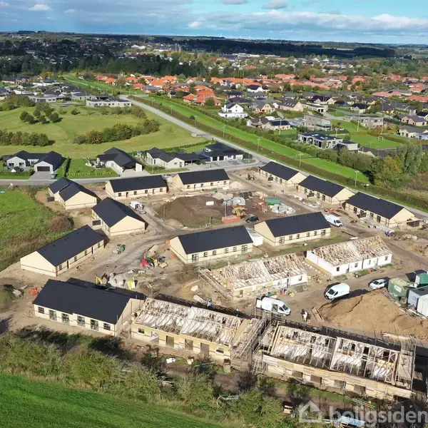 Byggeplads med række af nye huse under konstruktion; materialer og køretøjer spredt omkring. Beliggende i et forstadsområde med marker og flere boligområder i horisonten.