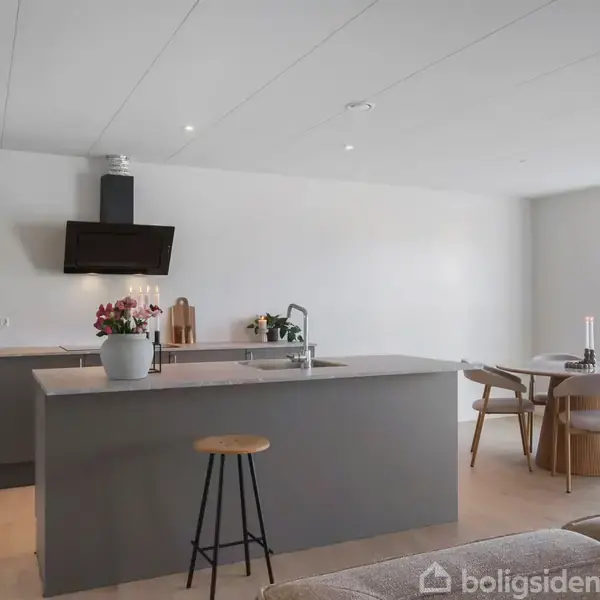 Køkkenø med blomster står i moderne køkken. Der er indsatser, herunder en vandhane og barstol. I baggrunden ses et spisebord med stole ved et stort, lyst vindue.