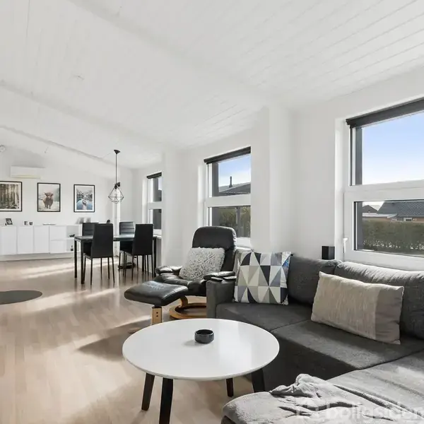 Grå sofa med puder står ved store vinduer i en lys, moderne stue med hvidt loft og trægulv. Spisebord og stole ses nær bagvæggen med dekorative billeder.