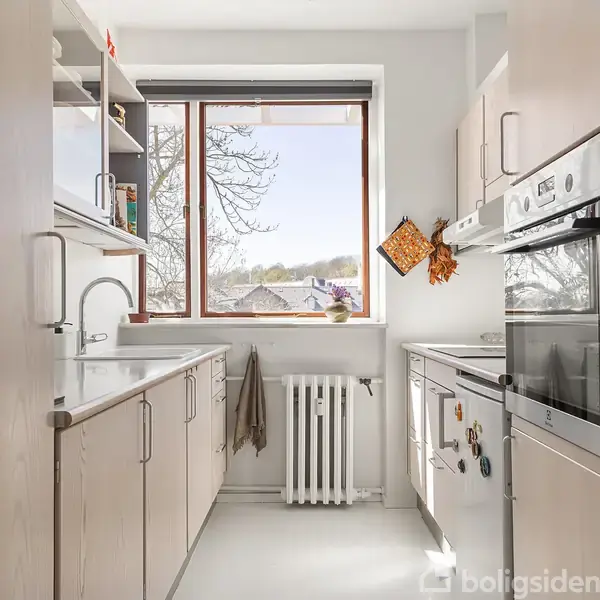 Køkken med lyse træskabe og moderne apparater. Sollys strømmer ind gennem vinduet, der viser et udsyn til et træ og huse udenfor. Hvid væg og radiator under vinduebænk.