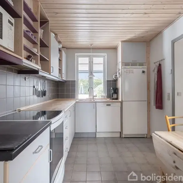 Et moderne køkken med hvide skabe og grå fliser. Der er en ovn med kogeplader, en mikrobølgeovn på hylden, og vinduet giver udsigt over grønne omgivelser. Bordet har stole omkring.