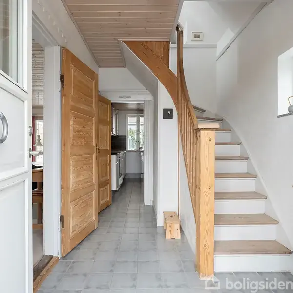 Trappe fører opad i en lys entré med hvidmalede vægge og trægulve. Åben dør til venstre giver kig til spisestue og køkken. Vindue til højre lader lys ind.