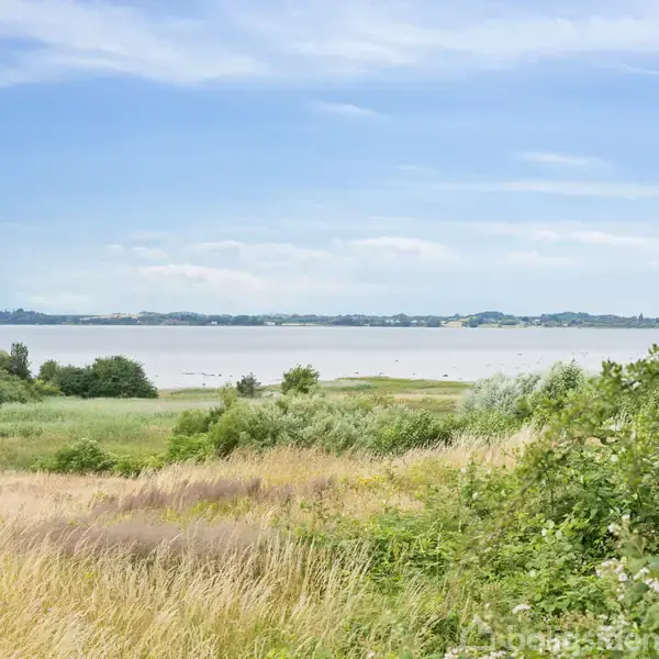 Et græsdækket sti fører mod en sø i baggrunden. Til højre står et lille halvtag, omgivet af højt græs og buske, under en klar blå himmel.