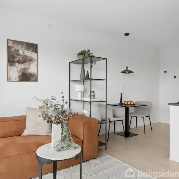 Brun sofa står stille i en moderne stue med et vægmaleri. Sort reol med pyntegenstande deler rummet, der også har et rundt spisebord med stole ved siden af et køkkenområde.