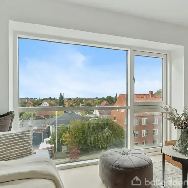 En lys stue med en stor vinduesfront, der viser en byudsigt med tætte huse og træer. En sofa og puf står foran vinduet, omgivet af dekorative puder og en vase.