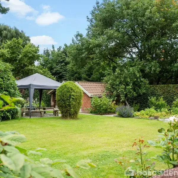 En have med en græsplæne og roser i forgrunden. En pavillon står på græsset med et bord under. I baggrunden ses et lille murstenshus omgivet af træer.