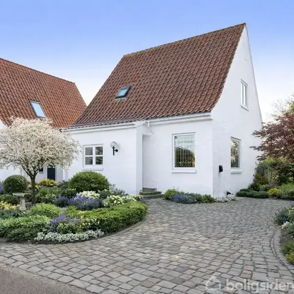 To hvidmalede huse med røde tegltage står omgivet af en velplejet have med blomster og træer. Foran husene ligger en brostensbelagt indkørsel.