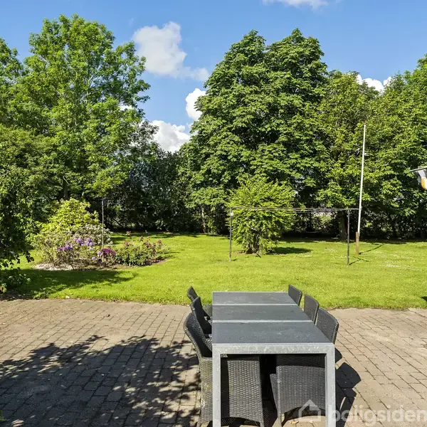 Et havesæt står på en flisebelagt terrasse. I baggrunden ses en stor græsplæne omgivet af frodige, grønne træer under en klar blå himmel.