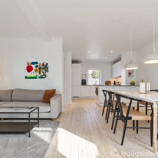 Stue med grå sofa og sofabord, åben spisestue med sort stole om hvidt bord i træ. Moderne lysindretning, køkken i baggrunden. Væggene har moderne kunstværker. Ingen tekst.