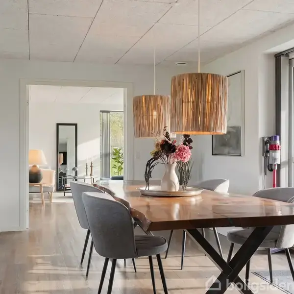 Et spisebord med en blomsterdekoration står i midten af et lyst rum. To pendellamper hænger over bordet. Store vinduer giver udsigt til en have. Ingen tekst bemærket.