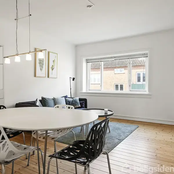 Et hvidt, rundt bord omgivet af designerstole står i en lys stue. I baggrunden ses en grå sofa foran en rektangulær vindue, med trægulv og moderne dekorationer omkring.
