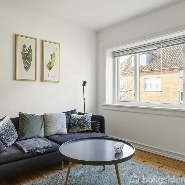 En mørk sofa med puder står ved et stort vindue, belyst af naturligt lys. To plantebilleder hænger på væggen over sofaen, og der er et rundt bord foran sofaen.
