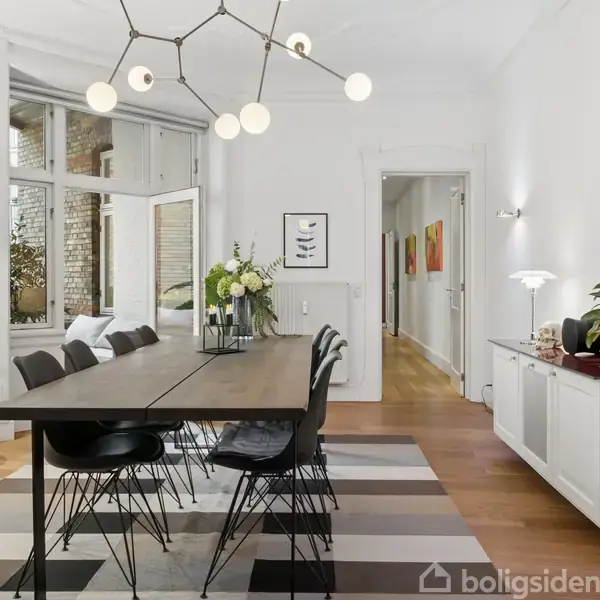 Spisebord med sorte stole står i en lys spisestue. Moderne lysekrone hænger over bordet. Langt hvide skab med planter til højre. Store vinduer indrammer baggrunden.
