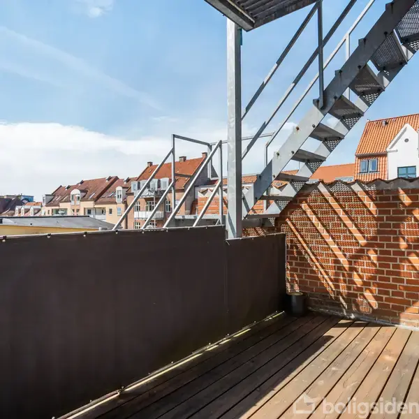 En trappe fører op fra en tagterrasse med trægulv og murstensvægge. Omkring ses byhuse med røde tage og en kirketårn i baggrunden under en blå himmel.