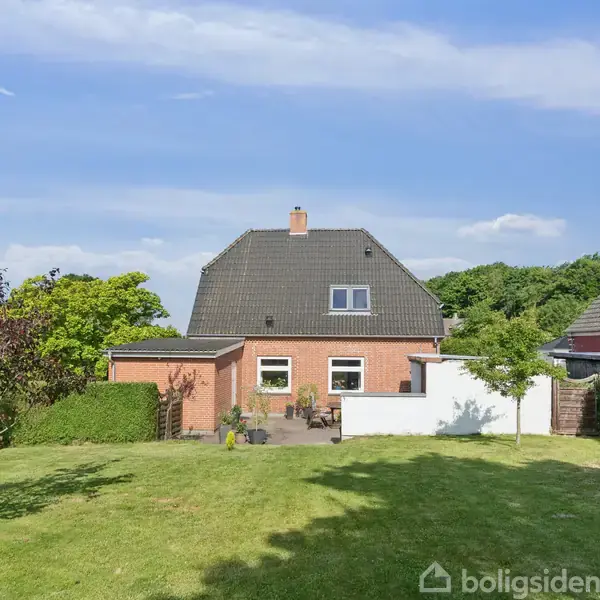 Et rødt murstenshus med gråt tag står i en velplejet have med græsplæne og træer under en blå himmel. Hvide vinduer ses på husets facade.