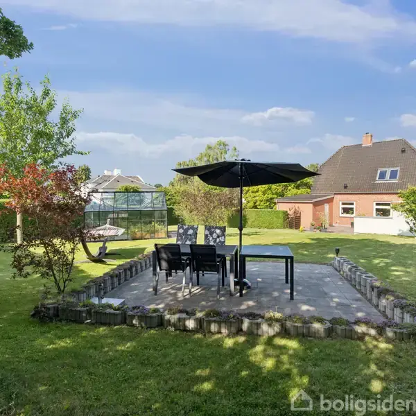 En terrasse med sorte havemøbler og en stor parasol står i en grøn have omkranset af en hæk. I baggrunden ses et hus med rødt tag.