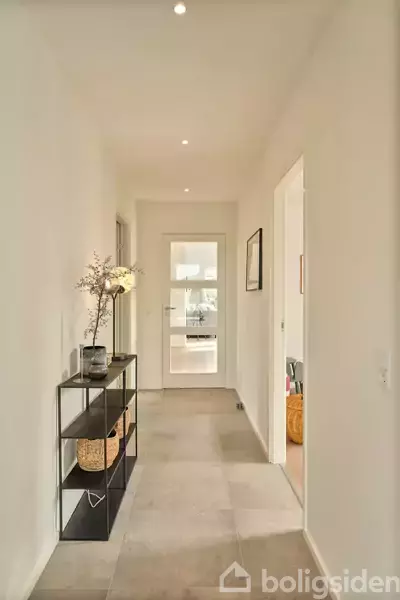 Gangen har en smal reol til venstre med dekorative genstande og en blomst. En dør i baggrunden leder til et andet rum. Væggene er hvide.