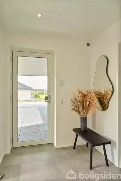 En smal entré med en glasdør til venstre åbner ind til en gårdhave. En skulpturel vægspejl hænger over en sort bænk med en vase fyldt med tørrede græsser.