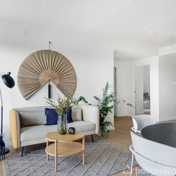 Sofa står i en stue, dekoreret med en stor vægvifte og planter. Et rundt sofabord med vaser og en læselampe ved siden af. Et spisebord med stole står i nærheden.