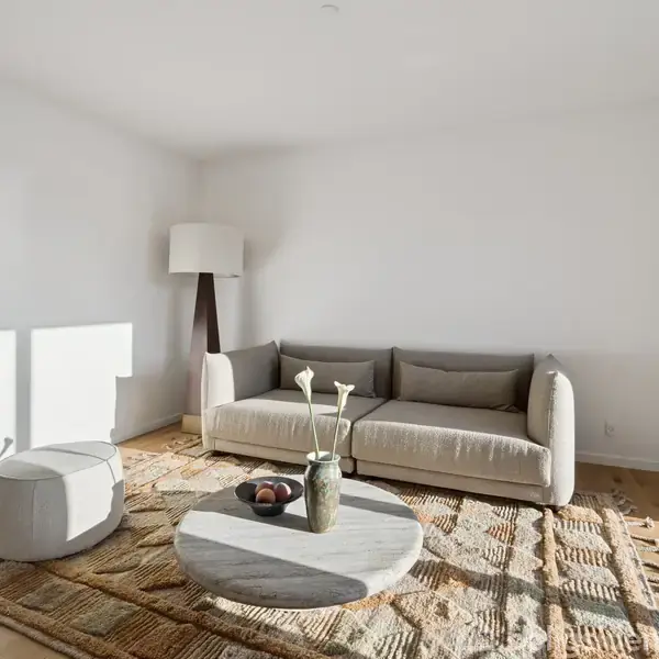 En lys stue med en beige sofa står mod en hvid væg. Foran står et rundt marmor sofabord med dekorative genstande. En gulvlampe står i hjørnet, og et mønstret tæppe dækker parketgulvet.