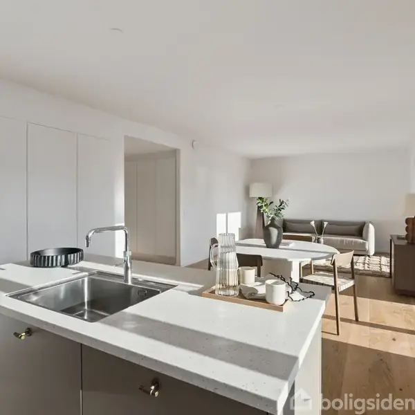 Køkkenø med vask står centralt i hvidt, lyst rum med sofa og bord i baggrunden. Store vinduer lader naturligt lys strømme ind.