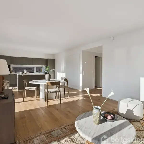 Lys stue med spisebord og stole centralt placeret. Rundt om ses et sofabord med dekorationer, en kommode med lampe, og et lyst køkken i baggrunden bag spiseområdet.