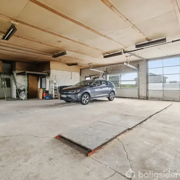 En bil holder parkeret i en rummelig garage med store glasporte og betonvægge. En åbnet værkstedsdør er synlig til venstre, og der hænger loftslamper.