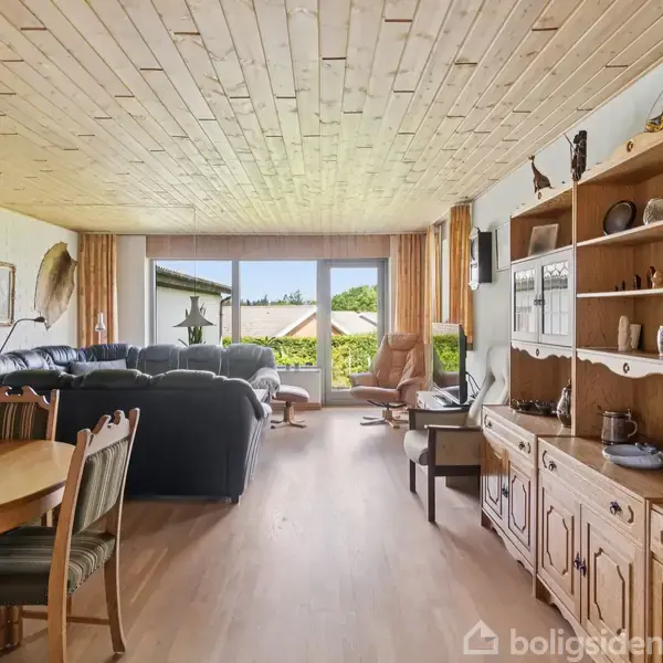 En stue med træbeklædt loft, en sort lædersofa ved vinduer med udsigt til en have. Der er et spisebord med stole og en stor reol med dekorative genstande.