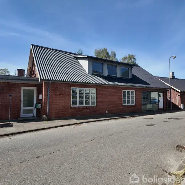 Rødstensbygning står i et gadehjørne med mosbegroet tag. Den har store vinduer og en glasdør i facaden. Fortovet er på højre side, mens der er buske bagved.