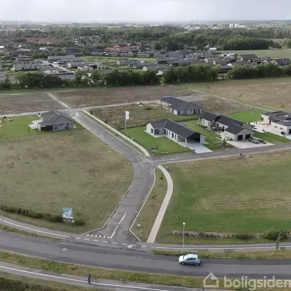 Huse står langs en vej i et åbent grønt område. En bil kører på en større vej i forgrunden. I baggrunden ses flere huse og grønne marker.