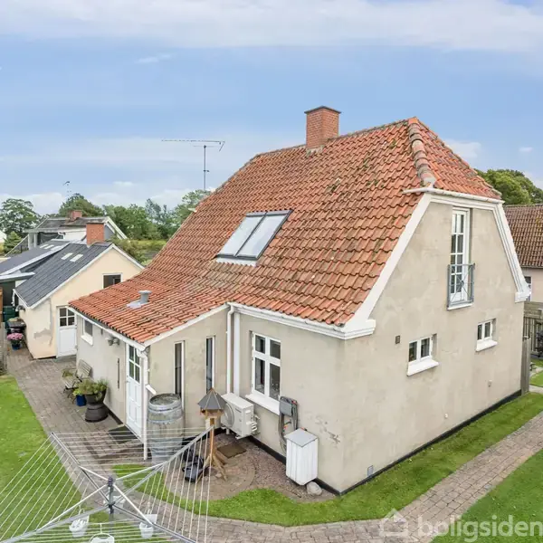 Et to-etagers hus med rødt tegltag står i en velplejet have. Solen skinner på terrassen, hvor der er planter og et tørrestativ. Omkring ligger andre huse i et villakvarter.