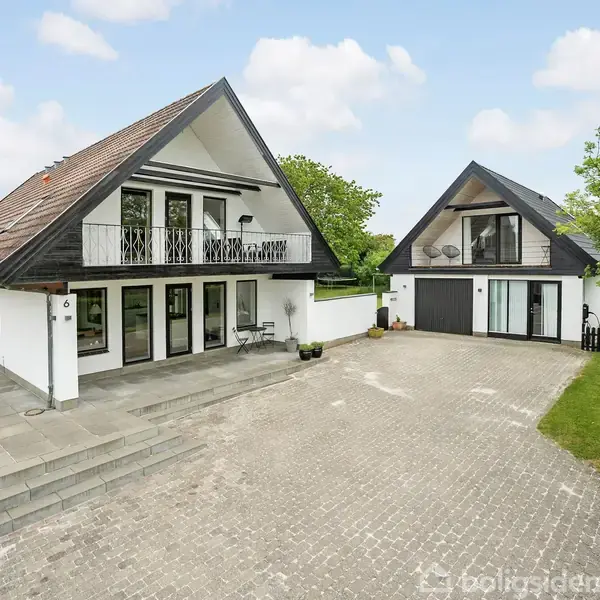 En hvid, to-etagers bygning med skråt tag og balkon står ved siden af en mindre bygning, der fungerer som garage. De er omgivet af grønne områder og belægningssten. Indgangen er nummereret "6".