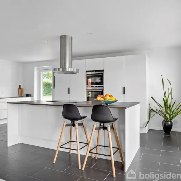 Køkken med hvidt køkkenbord knyttet til en ø, hvor to sorte barstole står. En frugtskål er på bordet, og en grøn plante står i hjørnet.