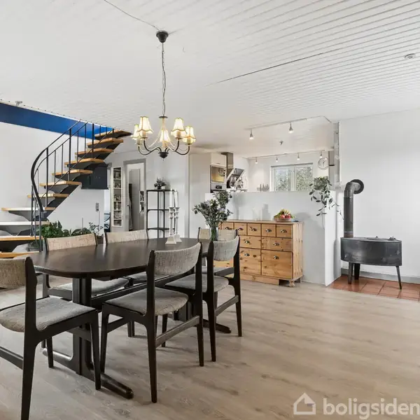 Spisebord står i stue med trægulv, omgivet af spisestole. En moderne trappe fører til overetagen. Ved siden af står en brændeovn. En kommode står nær et åbent køkkenområde.
