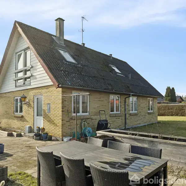 Et toetagers hus med gavltag står stille i en åben have. Foran huset er der en terrasse samt havemøbler og plantekrukker.