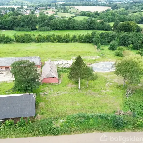Luftfoto af et landligt område med to bygninger og en cirkulær struktur omgivet af grønt græs og træer. Baggrunden viser vidstrakt skov og marker.