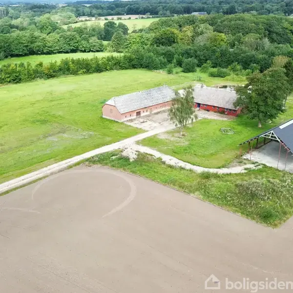 Et rødt hus med stråtag og et åbent skur står på en stor grøn mark. Omkring dem er der marker og skovområder i en landlig landskab.