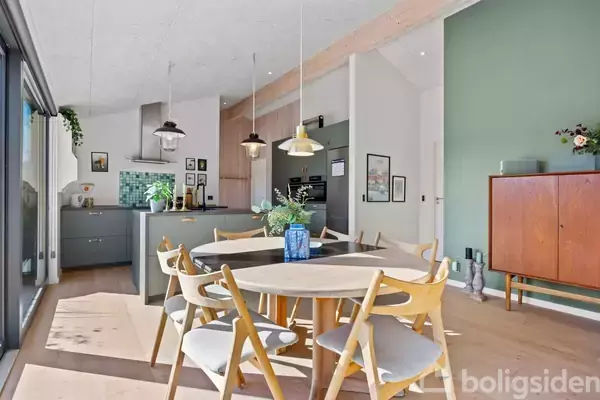 Rundt spisebord med træstole står i stuen. Over bordet hænger lamper, og bagved ses et åbent køkken med mintgrønne fliser. Væggene er grønne med kunstværker og skænk.