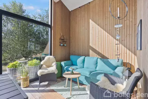 En lys stue med trævægge har en turkis sofa og to fletstole med fåreskindstæpper. Der er et rundt sofabord og planter. Store glasvinduer giver udsigt til træer udenfor.