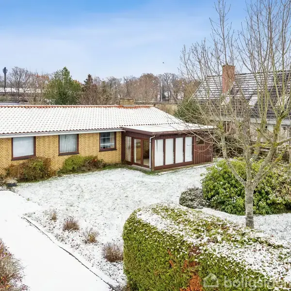 En etplans gulstensvilla med hvide tagsten står i en vinterhave med let snefyldt græsplæne og buske. Et nærliggende hus og træer er synlige i baggrunden.