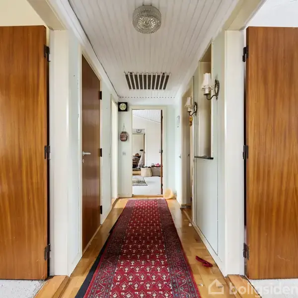 Gang med lange, røde tæpper, flankeret af trædøre på begge sider. En lampe hænger fra loftet, mens vægspejle og lanterner pynter væggene. En stue skimtes for enden.