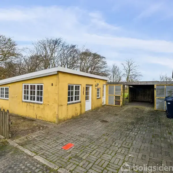 En gul bygning med hvide vinduer og dør står ved siden af en garage. Indkørslen er belagt med sten, omgivet af træer og buske.