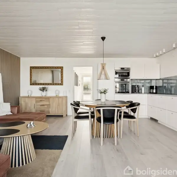En stue med brunt læderhjørnesofa, rundt sofabord, og spisebord med seks stole. Køkkenet har hvide skabe og en grå backsplash. Væggene er dekoreret med et spejl og en trækommode.