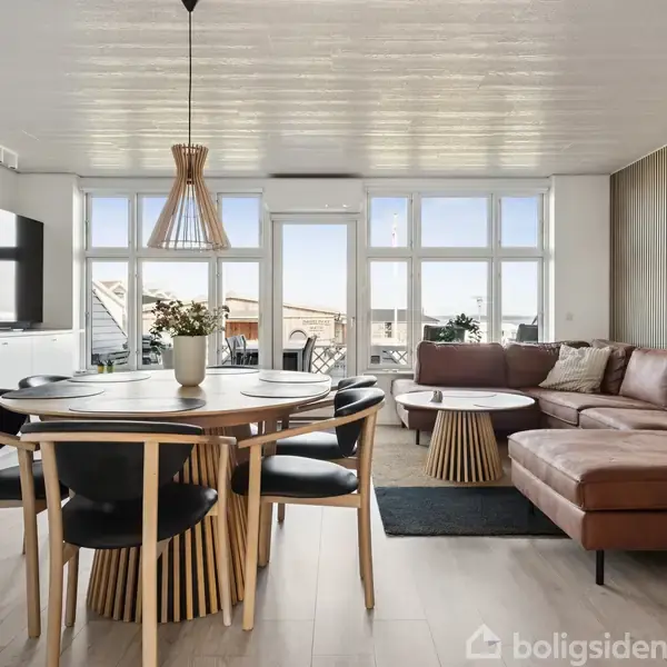 Spisestue med rundt bord og stole, der står i lys stue. Lædersofa i hjørnet, stort vindue bagved. TV monteret på væggen. Moderne indretning med trædetaljer.