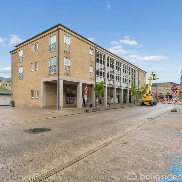Et tre-etagers gult murstensbygning står på en åben plads. En liftbil arbejder ved bygningen, og der er vejskilte i nærheden. Himlen er klar med få skyer.