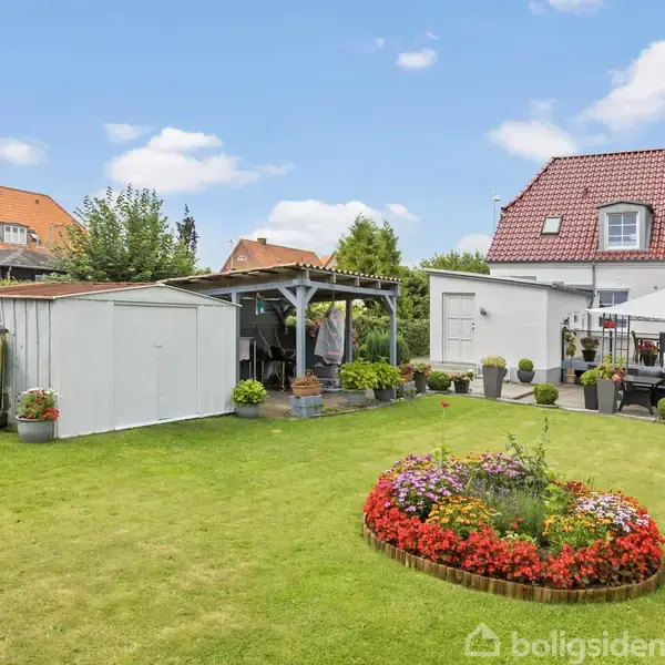 Græsplæne med rund blomsterbed i forgrunden; i baggrunden står et hvidt hus med rødt tag og et skur med halvtag, omgivet af havemøbler og krukker.