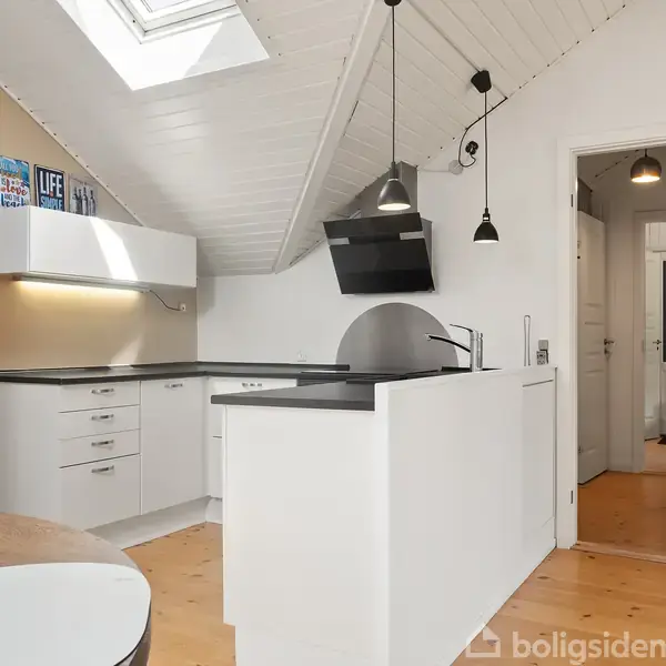 Et moderne køkken med hvide skabe og sort bordplade. Lamper hænger fra det skrå loft. Et lyst træbord med stole står i forgrunden. Skilttekst: "LIFE."