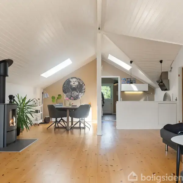 Brændeovn opvarmer en moderne stue med skrånende træloft. Et spisebord står ved siden af planten. Loftsvinduer lader lys ind, mens køkkenet ses i baggrunden.